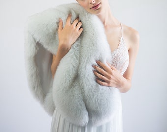 May include: A woman wearing a white dress and a white fur shawl. She is holding the shawl in front of her and looking at the camera. She is wearing a silver headband with small white flowers.