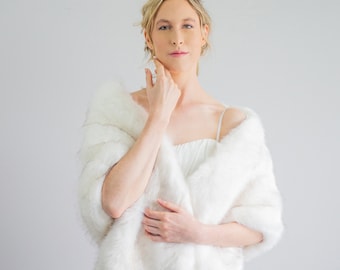 May include: A white faux fur shawl with a soft, fluffy texture. The shawl is draped over the shoulders of a person wearing a white dress.