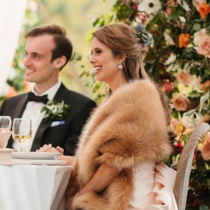 Puede incluir: Una novia y un novio están sentados en una mesa, la novia lleva una estola de piel sintética y un vestido blanco. El novio lleva un esmoquin negro con una pajarita blanca. Están sentados frente a un arreglo floral con una variedad de flores en tonos de rosa, naranja y blanco.