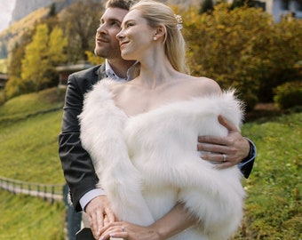 May include: A couple poses for a wedding photo in front of a waterfall and a mountain. The bride is wearing a white faux fur shawl and the groom is wearing a black suit.