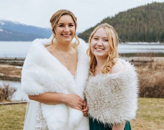 Puede incluir: Dos mujeres con chales de piel sintética. Una mujer lleva un chal blanco sobre un vestido de novia blanco con detalles de encaje. La otra mujer lleva un chal marrón claro sobre un vestido verde oscuro. Están sonriendo.