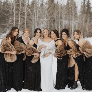 May include: A group of women in formal attire posing outdoors in a snowy setting. Most are wearing black gowns and faux fur wraps. One woman wears a white gown and a white faux fur wrap. The background features a forest.
