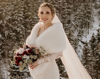 Puede incluir: Una novia con un vestido de novia de encaje blanco con un chal de piel blanca y un velo, sosteniendo un ramo de flores rojas y blancas. Está de pie en un entorno de bosque nevado.