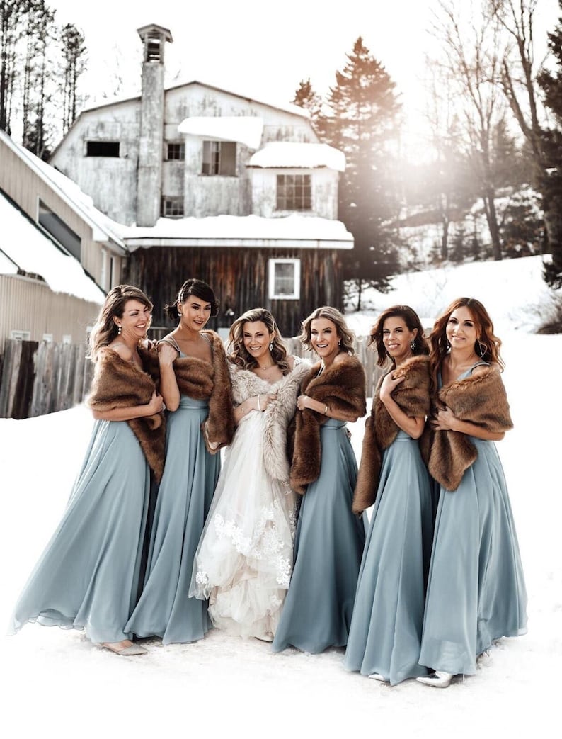 May include: Six bridesmaids in light blue floor-length dresses with brown fur wraps stand in a snowy outdoor setting with a white building in the background. The bride is in the center wearing a white lace wedding dress.