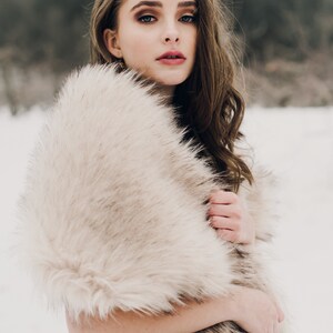 May include: A woman wearing a long, light brown faux fur shawl. She is standing in a snowy landscape.