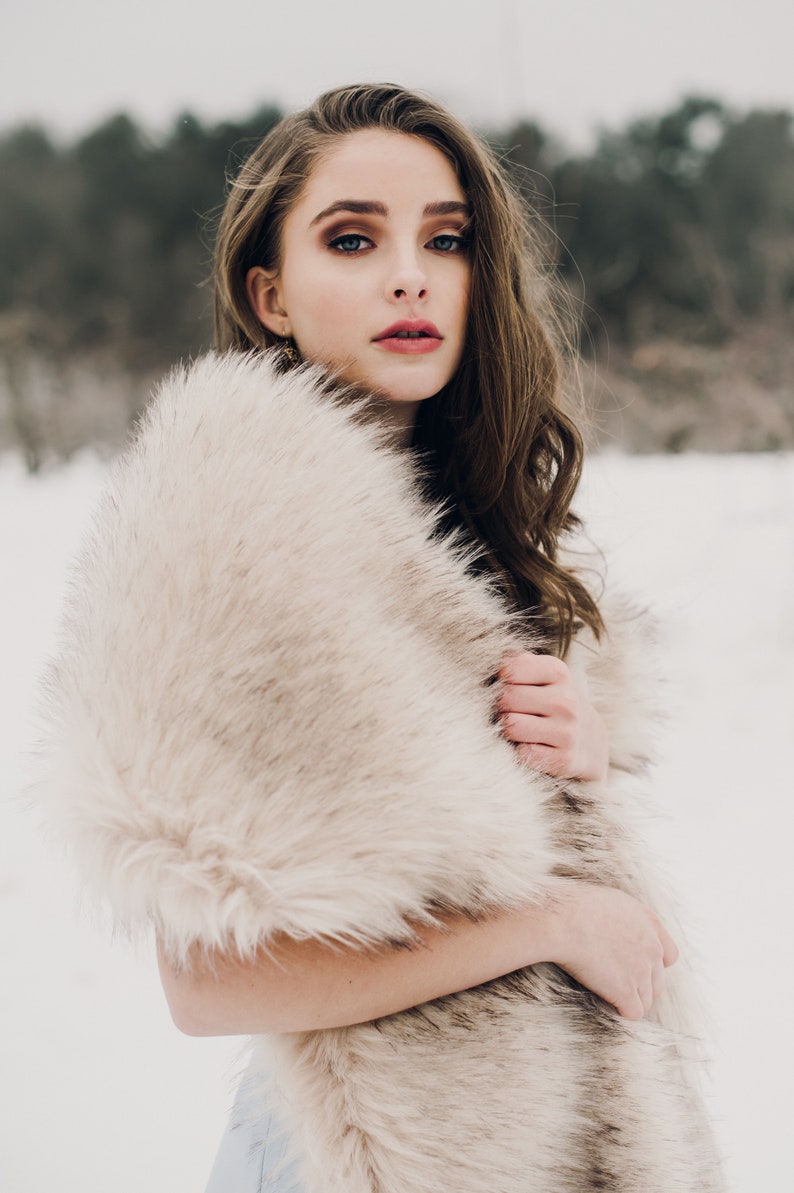 May include: A woman wearing a long, light brown faux fur shawl. She is standing in a snowy landscape.
