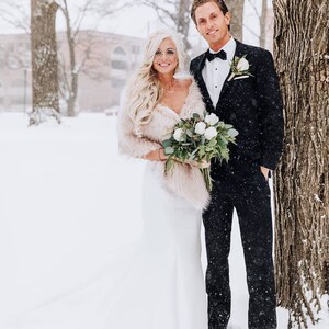 May include: A bride and groom pose for a wedding photo in a snowy outdoor setting. The bride is wearing a white wedding dress with a fur shawl and a veil. The groom is wearing a black tuxedo with a white bow tie. They are holding a bouquet of white flowers.