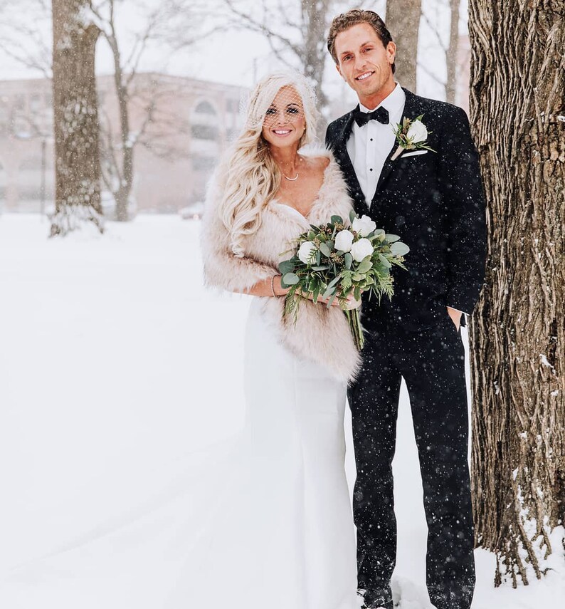 May include: A bride and groom pose for a wedding photo in a snowy outdoor setting. The bride is wearing a white wedding dress with a fur shawl and a veil. The groom is wearing a black tuxedo with a white bow tie. They are holding a bouquet of white flowers.