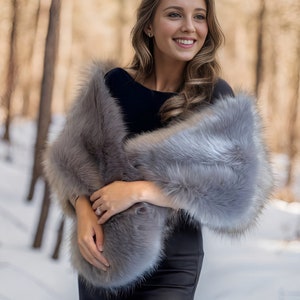May include: A woman wearing a gray faux fur shawl over a black dress. She is standing in a snowy forest with trees in the background.