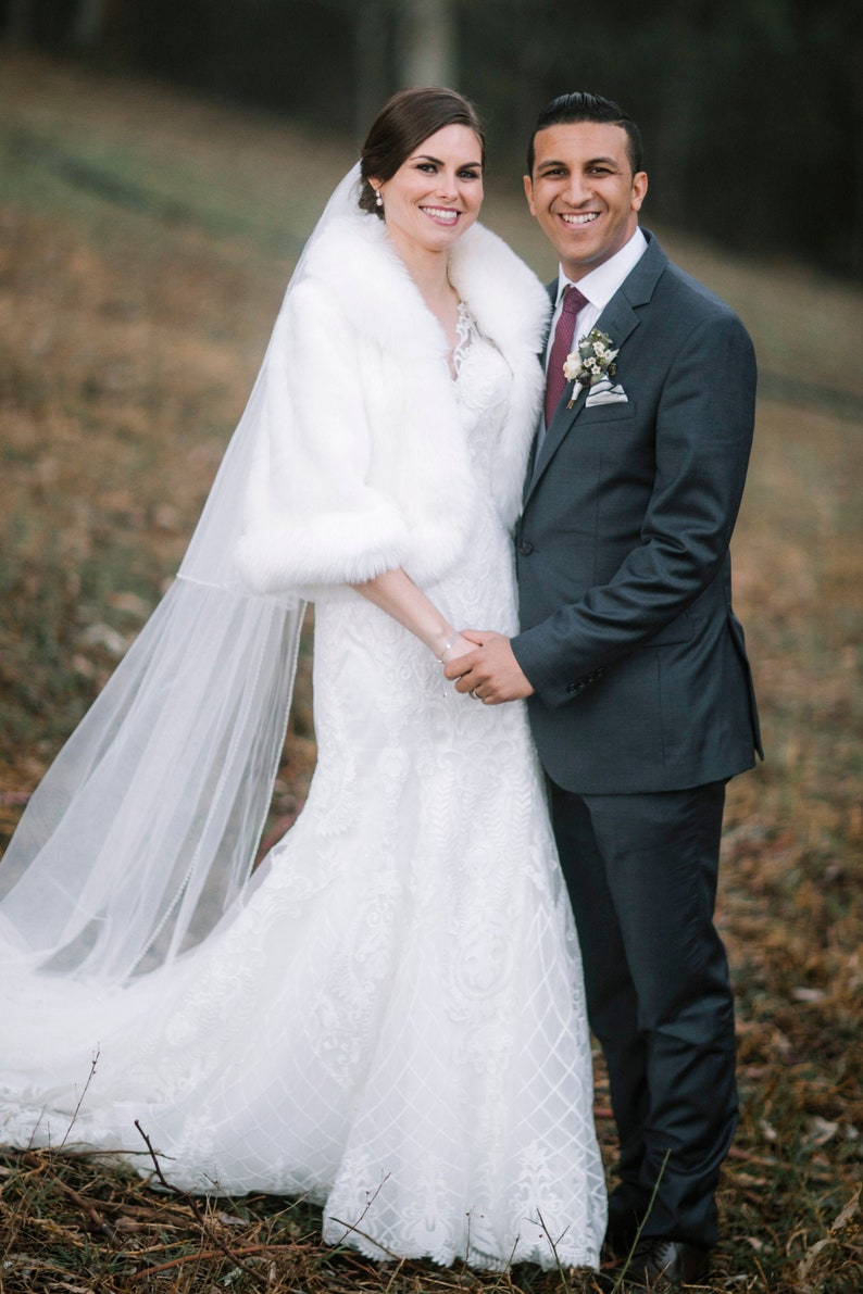 May include: A bride and groom pose for a wedding portrait. The bride is wearing a white wedding dress with a lace pattern and a white fur shawl. She is also wearing a veil. The groom is wearing a dark suit with a burgundy tie and a white shirt. They are standing in a field with brown grass and trees in the background.