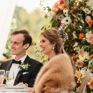 May include: A bride and groom sit at a table with a white tablecloth. The bride is wearing a white dress and a brown fur shawl. The groom is wearing a black tuxedo with a white bow tie. They are both smiling and looking at each other. There is a floral arrangement behind them.