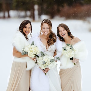 May include: Three women in winter attire stand in a snowy field. The woman in the middle is wearing a white wedding dress and a white fur shawl. The women on either side are wearing beige dresses and white fur shawls. All three women are holding bouquets of white flowers.