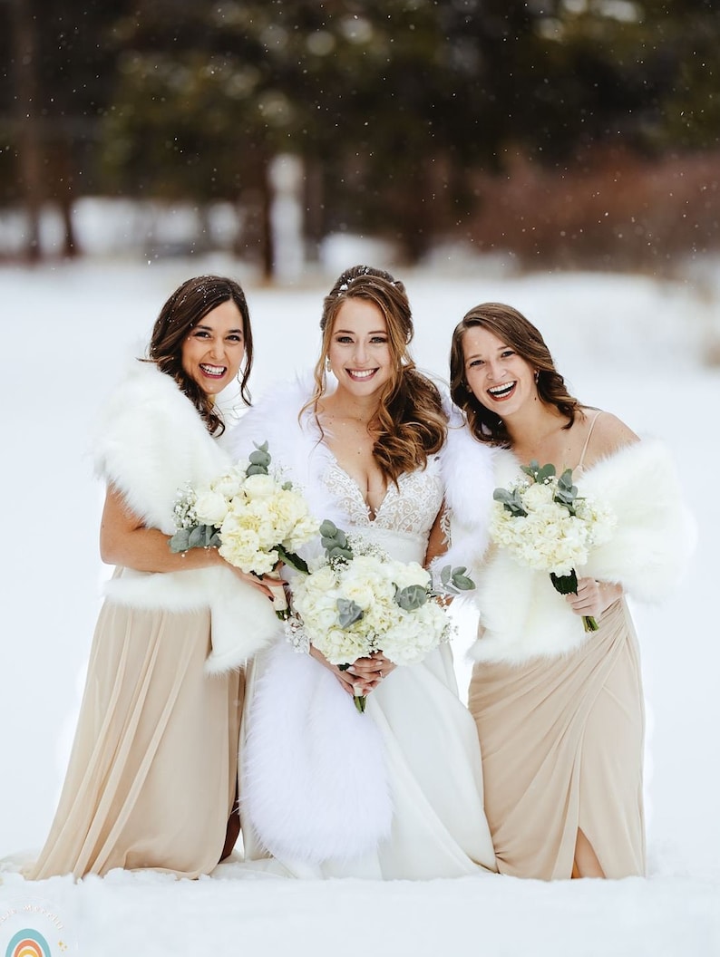 May include: Three women in winter attire stand in a snowy field. The woman in the middle is wearing a white wedding dress and a white fur shawl. The women on either side are wearing beige dresses and white fur shawls. All three women are holding bouquets of white flowers.