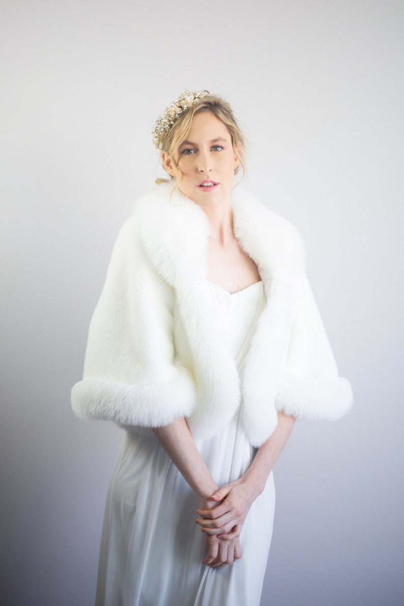 May include: A white faux fur bridal wrap with a wide collar and long sleeves. The wrap is made of soft, fluffy faux fur and has a luxurious feel.