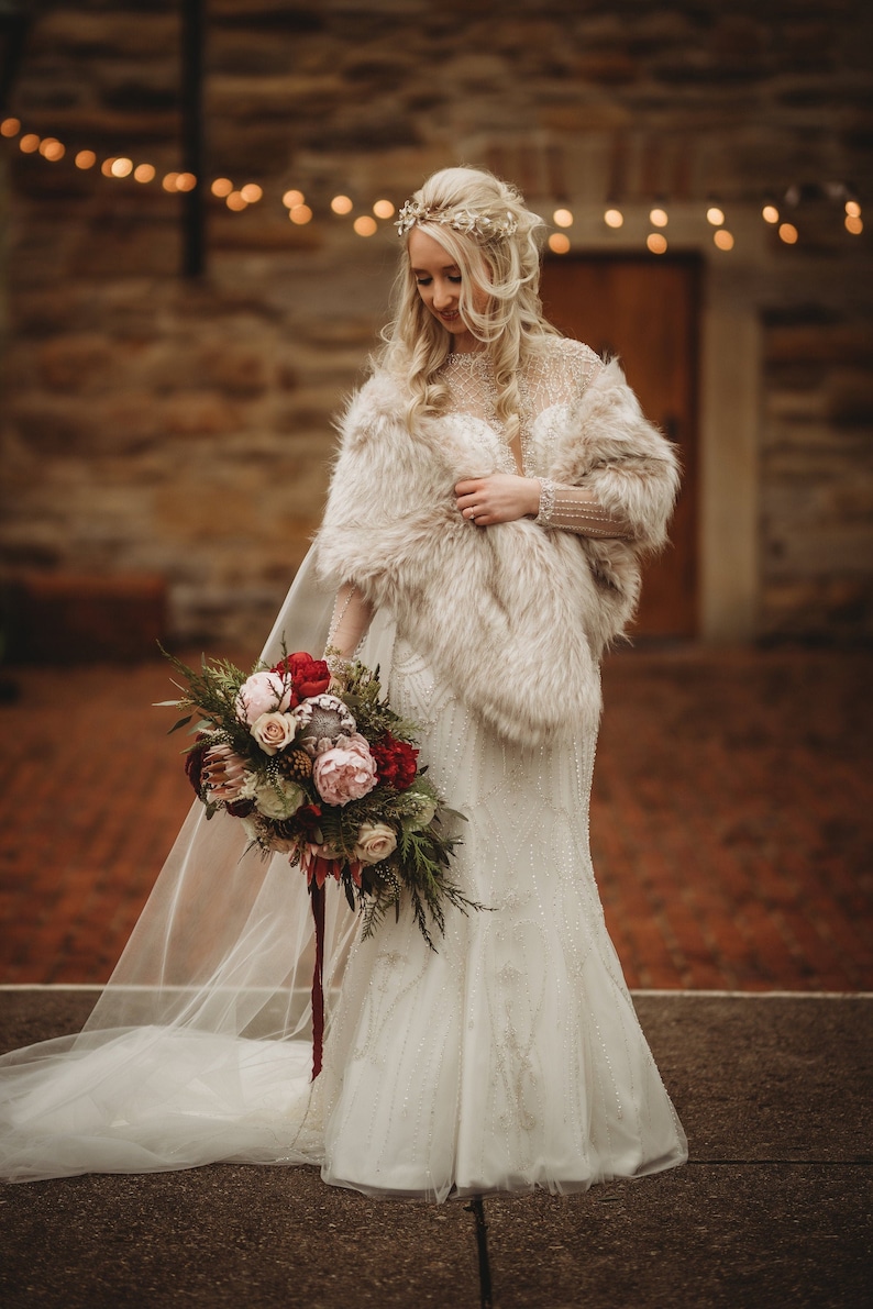 May include: A bride wearing a white wedding dress with a fur shawl and a bouquet of red, pink, and white flowers. She is standing in front of a stone building with string lights.