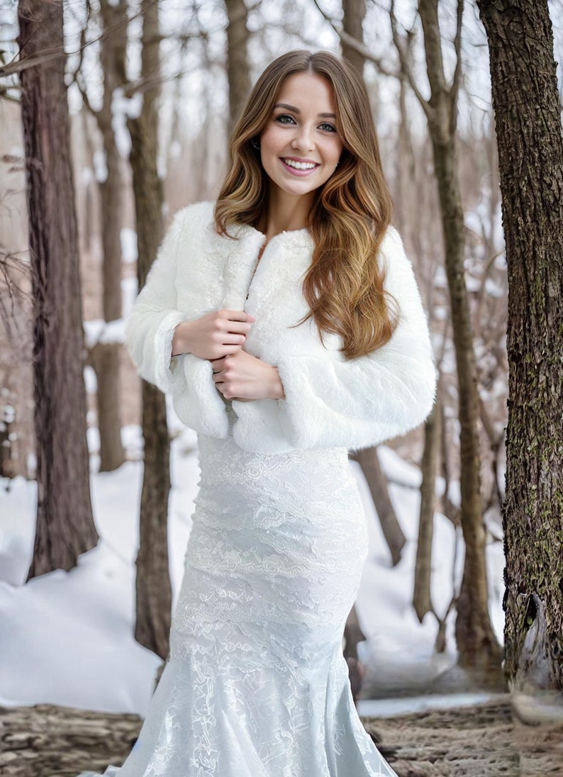 May include: A woman wearing a white faux fur shrug over a white lace dress. She is standing in a snowy forest with trees in the background.