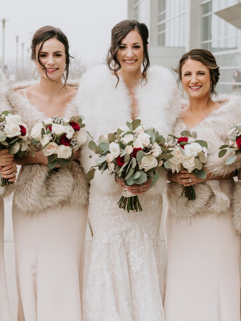May include: Three women wearing white faux fur wraps and holding bouquets of white and red roses. The woman in the middle is wearing a white wedding dress.