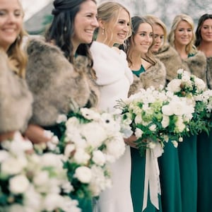 May include: A group of bridesmaids wearing teal green dresses and faux fur wraps stand with the bride in a white dress. They are holding bouquets of white flowers with green foliage.