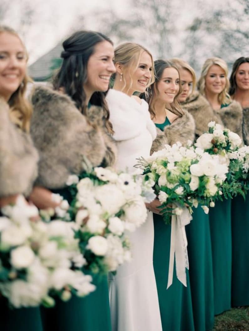 May include: A group of bridesmaids wearing teal green dresses and faux fur wraps stand with the bride in a white dress. They are holding bouquets of white flowers with green foliage.
