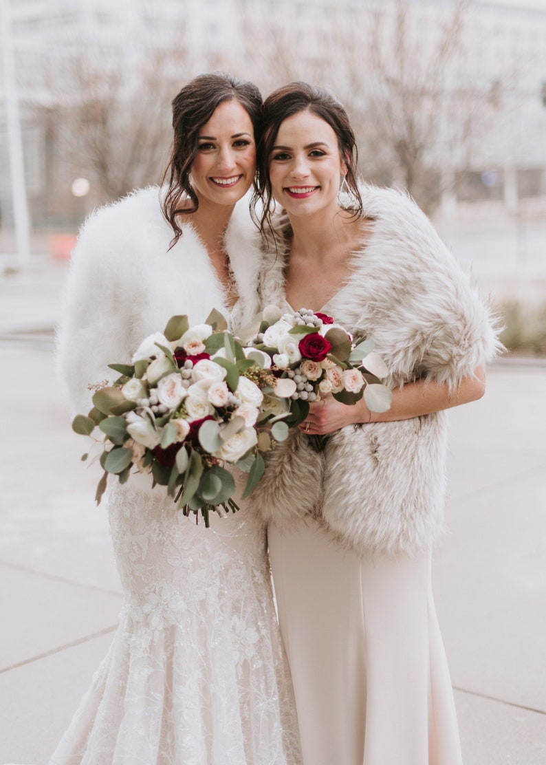 May include: Two people wearing white faux fur wraps and holding bouquets of white and red flowers. The person on the left is wearing a white lace wedding dress. The person on the right is wearing a blush pink dress.