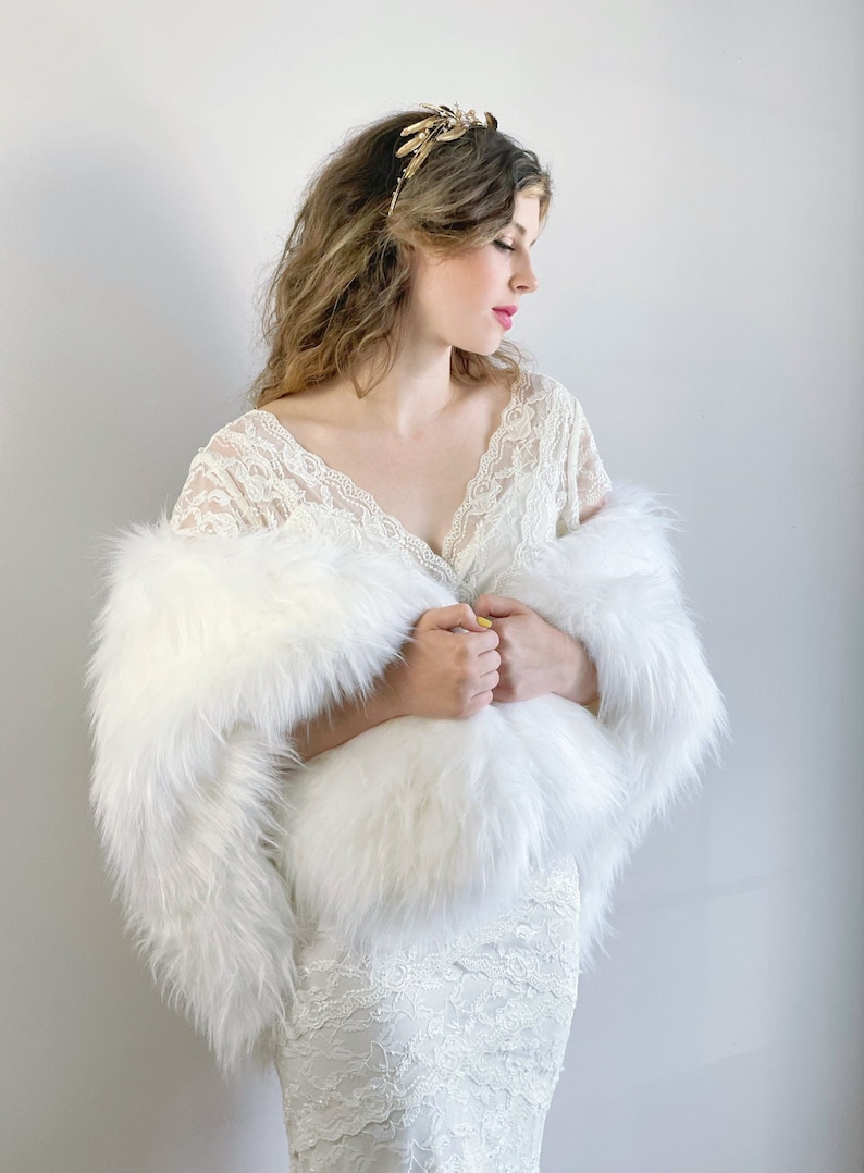 May include: A white faux fur shawl with a long, flowing design. The shawl is draped over a person wearing a white lace dress and a gold leaf headpiece.