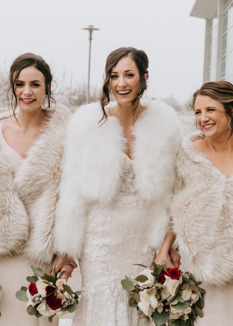 May include: Three women wearing white and cream faux fur shrugs, holding bouquets of white and red roses. The bride in the center is wearing a white lace wedding dress.