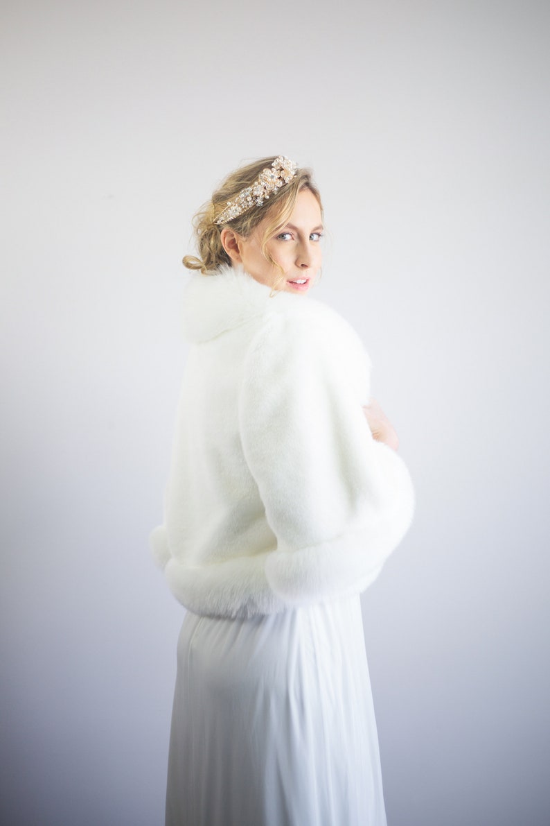 May include: A white faux fur bridal bolero with a long sleeve and a fur trim. The bolero is worn over a white dress.