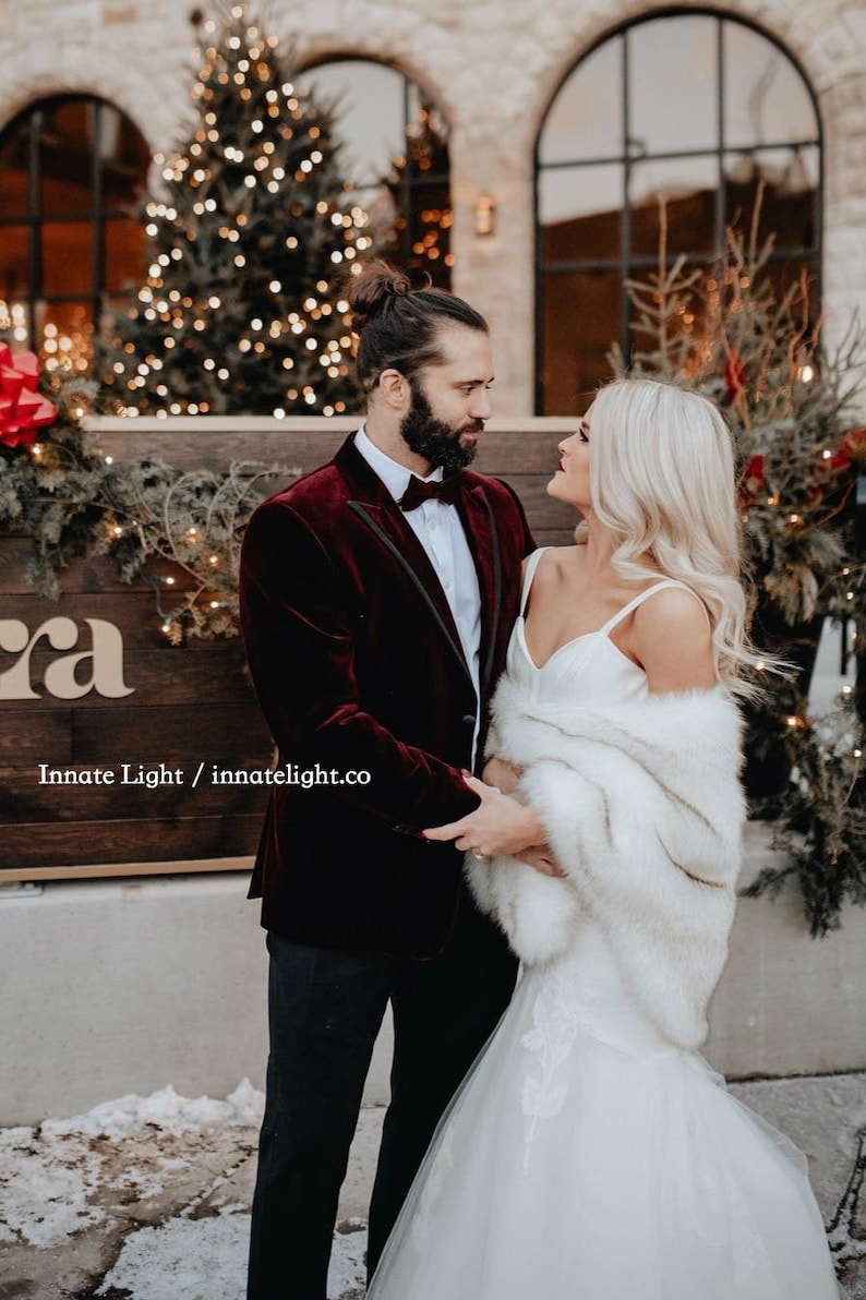 May include: A couple poses for a wedding photo in front of a Christmas tree. The groom is wearing a burgundy velvet suit jacket with a black bow tie. The bride is wearing a white wedding dress with a white fur shawl. The sign behind them reads "Innate Light / innatelight.co"