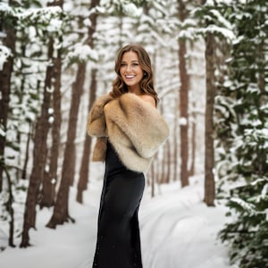 May include: A woman in a black dress and a brown fur shawl stands in a snowy forest. She is smiling and looking at the camera.