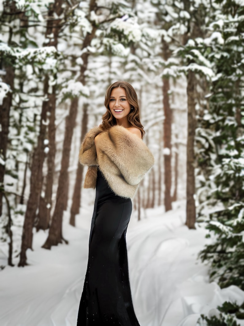 May include: A woman in a black dress and a brown fur shawl stands in a snowy forest. She is smiling and looking at the camera.