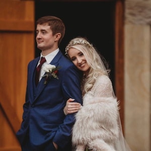 May include: A bride and groom pose for a wedding portrait. The bride is wearing a white dress with a fur shawl and a silver tiara. The groom is wearing a blue suit with a white shirt and a red tie. They are standing in front of a wooden door with string lights above.