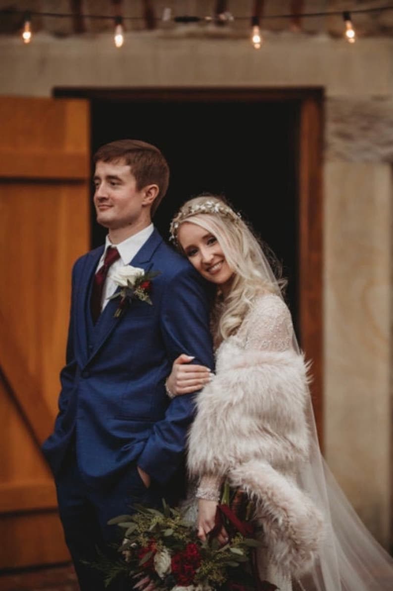 May include: A bride and groom pose for a wedding portrait. The bride is wearing a white dress with a fur shawl and a silver tiara. The groom is wearing a blue suit with a white shirt and a red tie. They are standing in front of a wooden door with string lights above.