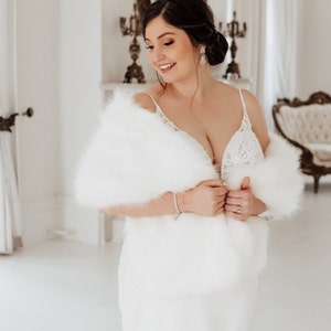 May include: A bride wearing a white wedding dress with a white fur shawl. She is standing in a white room with a mirror and a chandelier in the background.