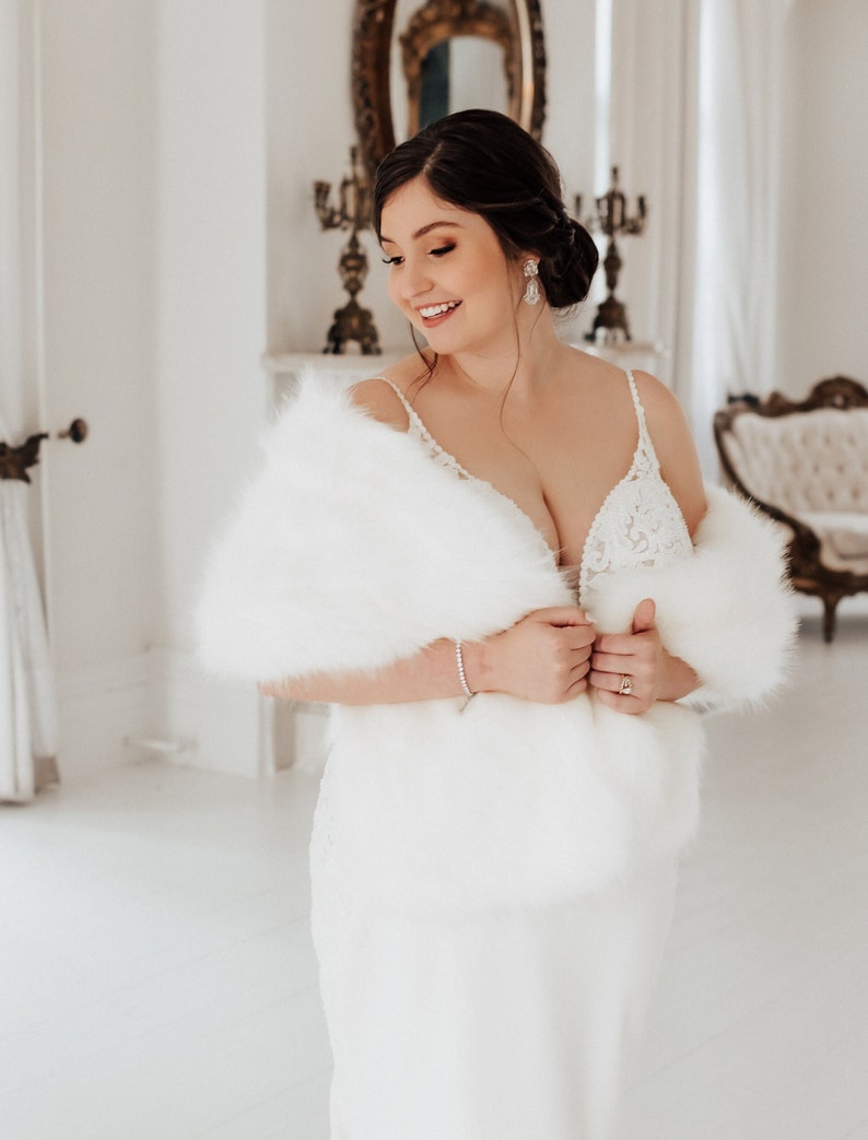 May include: A bride wearing a white wedding dress with a white fur shawl. She is standing in a white room with a mirror and a chandelier in the background.