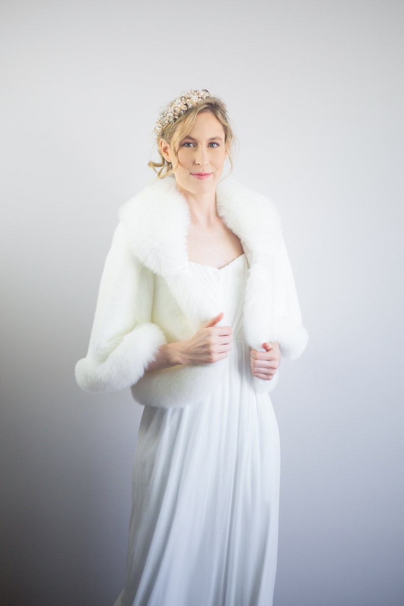 May include: A white faux fur bridal shrug with a large collar and long sleeves. The shrug is worn over a white dress.