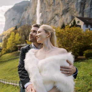 May include: A couple in wedding attire stand in front of a waterfall and a mountain. The bride is wearing a white faux fur shawl and the groom is wearing a black suit. They are looking up at the waterfall.