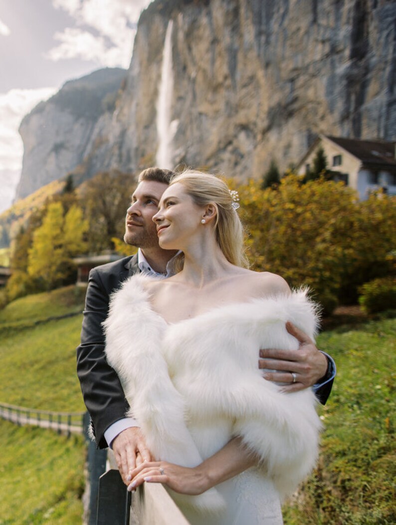 May include: A couple in wedding attire stand in front of a waterfall and a mountain. The bride is wearing a white faux fur shawl and the groom is wearing a black suit. They are looking up at the waterfall.