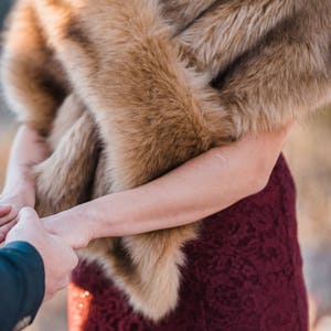 May include: A woman wearing a burgundy lace dress with a brown faux fur shawl draped over her shoulders. She is holding hands with someone off-camera.