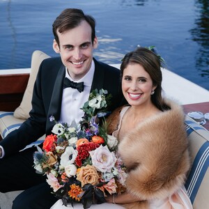 May include: A bride and groom pose for a photo on a boat. The bride is wearing a white dress with a fur shawl and holding a bouquet of colorful flowers. The groom is wearing a black tuxedo with a white shirt and black bow tie.