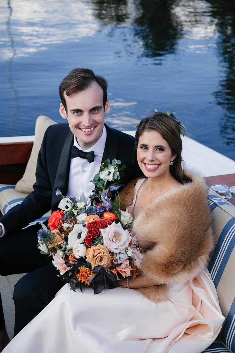 May include: A bride and groom pose for a photo on a boat. The bride is wearing a white dress with a fur shawl and holding a bouquet of colorful flowers. The groom is wearing a black tuxedo with a white shirt and black bow tie.