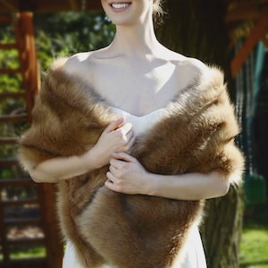 May include: A woman wearing a white dress with a brown faux fur shawl draped around her shoulders. She is standing in front of a wooden structure with green foliage in the background.