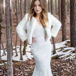 May include: A white faux fur bridal jacket with long sleeves and a cropped length. The jacket is worn over a white lace wedding dress with a fitted bodice and a flowing skirt.
