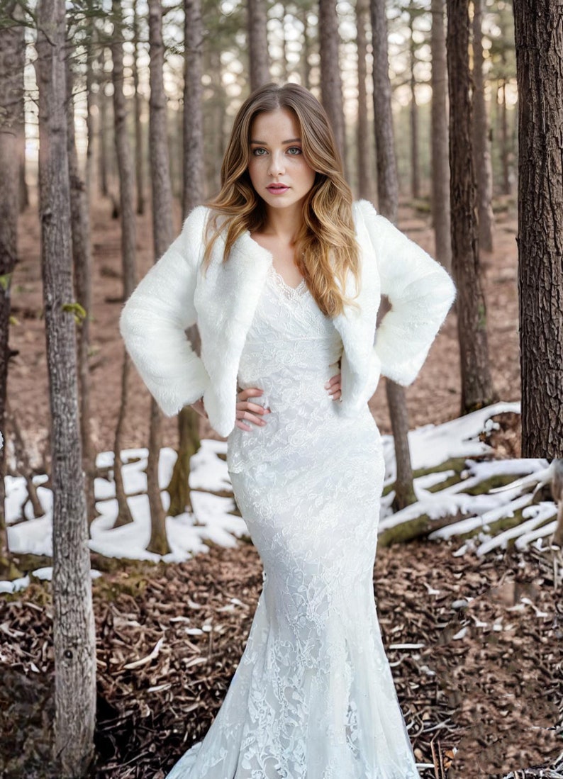 May include: A white faux fur bridal jacket with long sleeves and a cropped length. The jacket is worn over a white lace wedding dress with a fitted bodice and a flowing skirt.