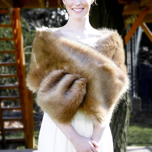 May include: A woman wearing a white dress with a brown faux fur shawl draped over her shoulders. She is standing in front of a wooden structure.