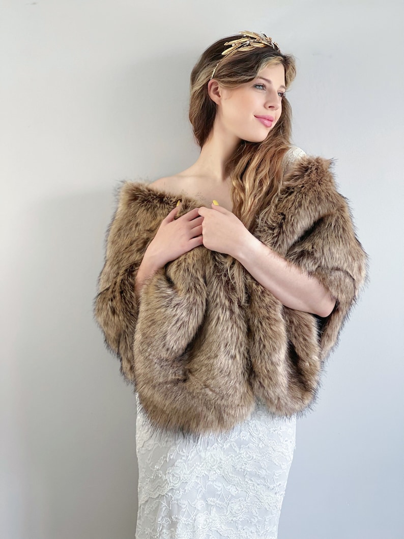 May include: A brown faux fur shawl with a long, flowing design. The shawl is draped over the shoulders of a person wearing a white lace dress.