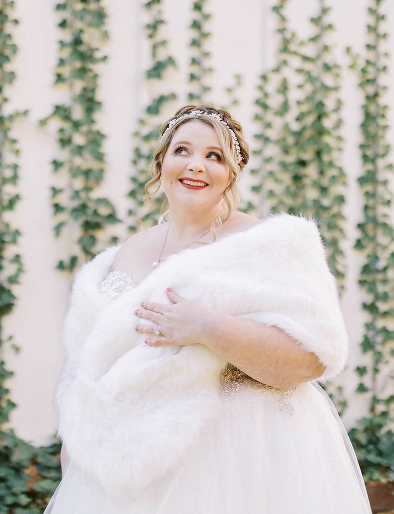 May include: A bride wearing a white wedding dress with a white fur shawl. She is smiling and looking up. The background is a white wall with green vines.