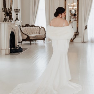 May include: A bride wearing a white wedding dress with a long train and a white faux fur shawl. She is standing in a white room with a fireplace and a chandelier.