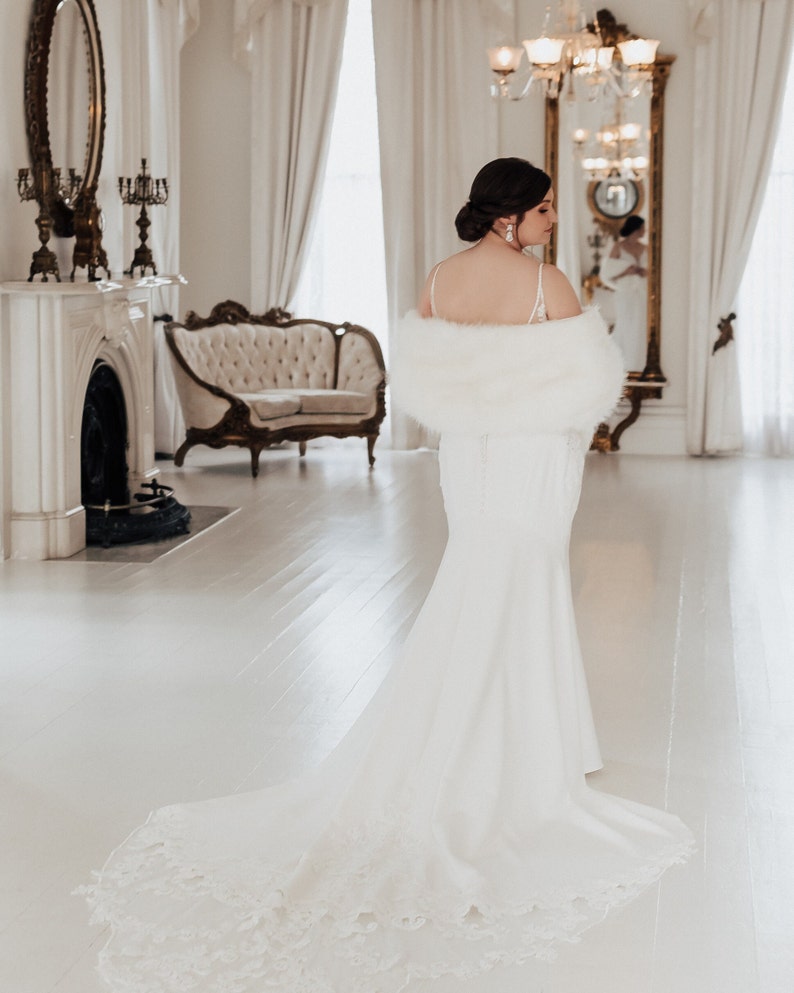 May include: A bride wearing a white wedding dress with a long train and a white faux fur shawl. She is standing in a white room with a fireplace and a chandelier.