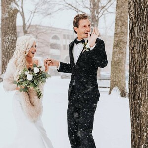 May include: A bride and groom in a snowy winter setting. The bride is wearing a white dress with a fur shawl and a bouquet of white flowers. The groom is wearing a black suit with a white shirt and a black bow tie. They are smiling and holding hands.