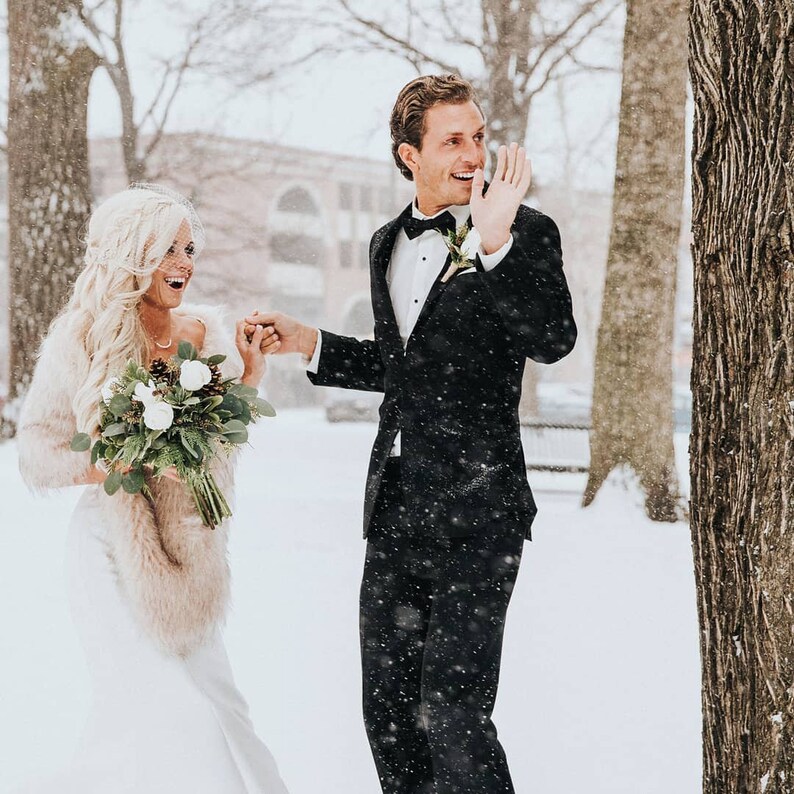 May include: A bride and groom in a snowy winter setting. The bride is wearing a white dress with a fur shawl and a bouquet of white flowers. The groom is wearing a black suit with a white shirt and a black bow tie. They are smiling and holding hands.